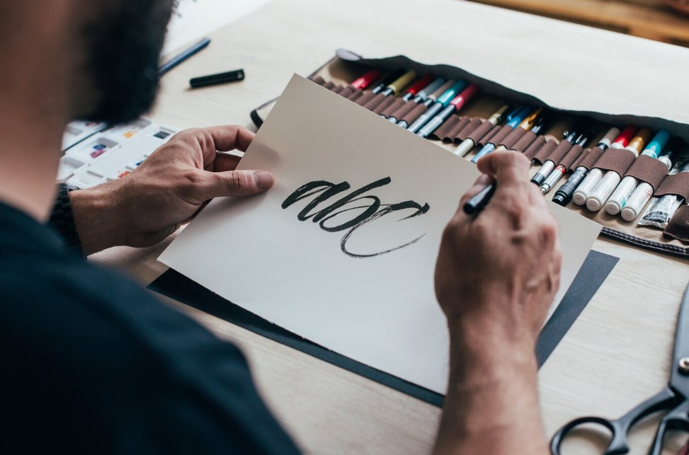 a bearded male designer holding a paper sheet with a hand-lettering saying “abc”