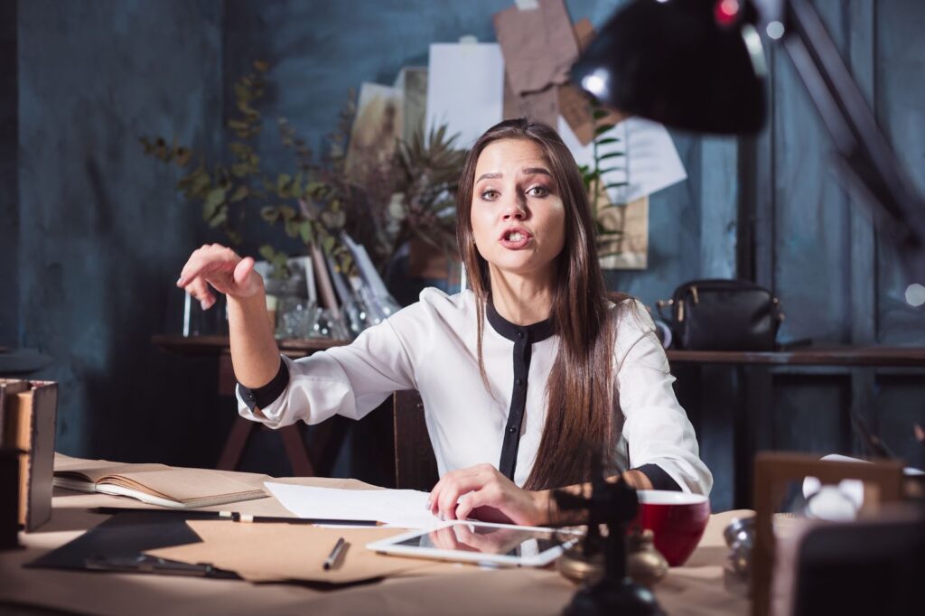 Woman sitting at a desk cluttered with papers who appears to be explaining something